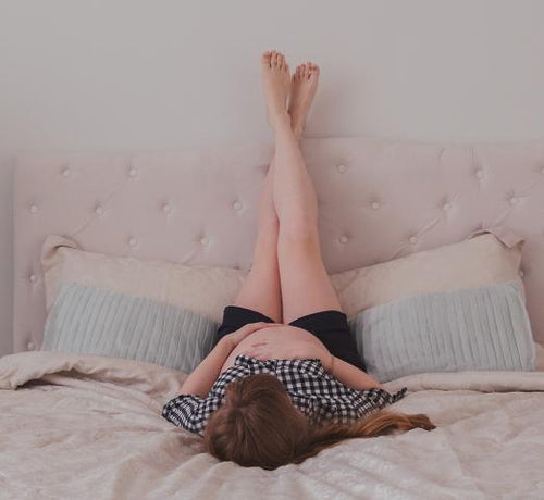 Femme enceinte allongée sur un lit rose avec les jambes levées contre le mur - Mustela Belgium - 1