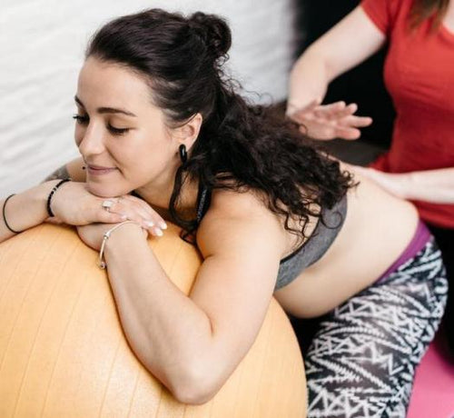 Femme enceinte appuyée contre un ballon de fitness se faisant masser le dos par une autre femme - Mustela Belgium - 1