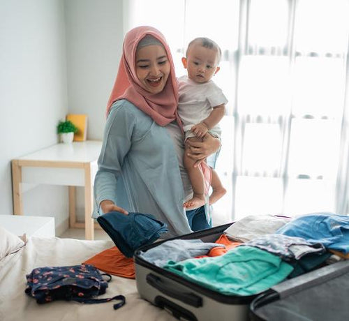 Maman qui tient son bébé dans ses bras et qui prépare une valise en mettant des vêtements à l'intérieur - Mustela Belgium - 1