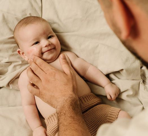 Bébé allongé sur un lot avec son père qui lui applique de la crème sur le visage - Mustela Belgium - 1