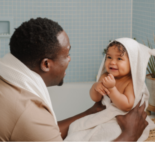 Bébé métisse avec une cape de bain qui sort du bain et qui est porté par son papa - Mustela Benelux - 1