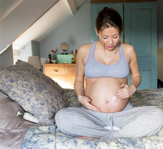Femme enceinte assise sur un lit qui applique de la crème sur son ventre