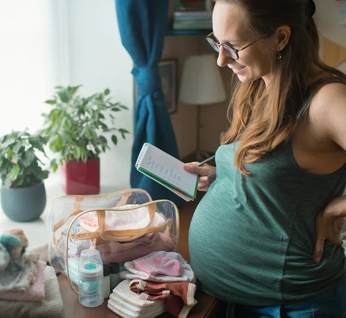 Femme enceinte faisant sa liste de valise maternité, carnet en main et main posée sur le ventre. - Mustela Belgium - 1