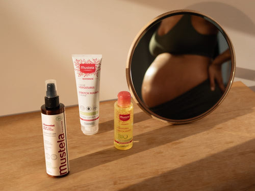 Produits de la gamme Maternité de chez Mustela posé sur une table, avec en fond le reflet dans un miroir d'une femme enceinte - Mustela Belgium - 4