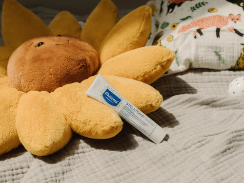 Crème Stelatopia posée sur un lit, entourée de coussins, dont un en forme de fleur - Mustela Belgium - 4