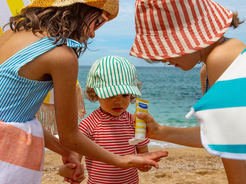 Enfant avec un bob à rayures sur la plage appliquant de la crème solaire dans sa main - Mustela Belgium - 2