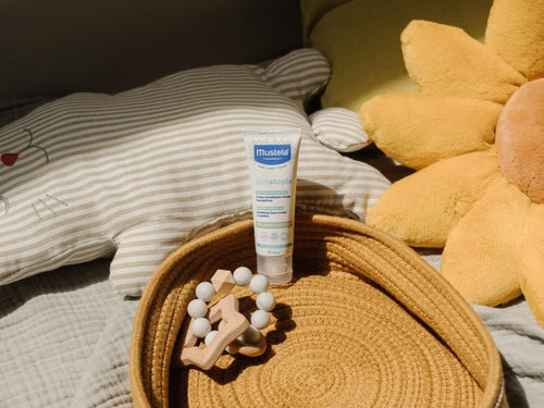 Stelatopia Crème émolliente pour le visage de Mustela posée sur un lit, entourée des affaires de bébé et de coussins - Mustela Belgium - 5